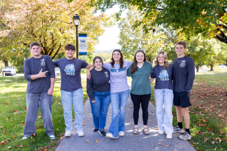 A group of seven diverse students stand in a row with their arms around each other's shoulders, smiling on a paved campus walkway. Lush green and autumn-colored trees line the path behind them.