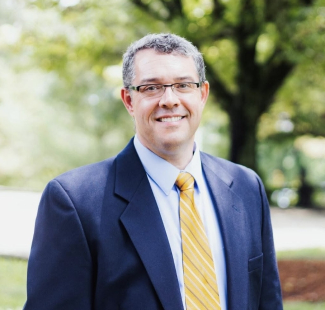 Andy Olson smiling outdoors, wearing a navy blue blazer, light blue shirt, and a yellow striped tie.