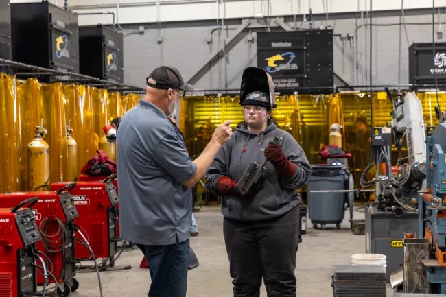 A welding instructor speaks to a student holding a metal piece in an industrial training shop. The student wears protective gear, including a raised welding helmet and heavy gloves, surrounded by red Lincoln Electric welding machines and yellow safety curtains.