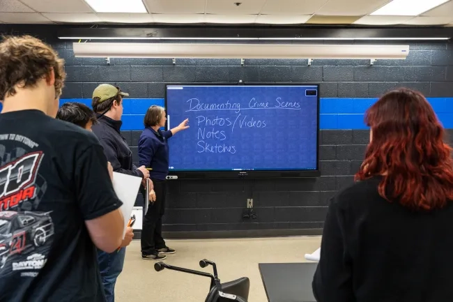An instructor points to a digital smartboard displaying handwritten notes on "Documenting Crime Scenes" while criminal justice students watch from the foreground in a classroom with a blue line painted on a dark brick wall.