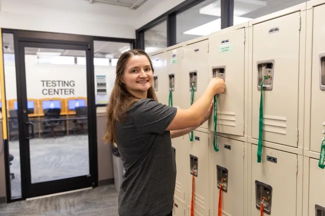A young woman with long brown hair smiles while opening a beige locker using a green lanyard key. In the background, a glass door reads "TESTING CENTER" with computer stations visible inside.